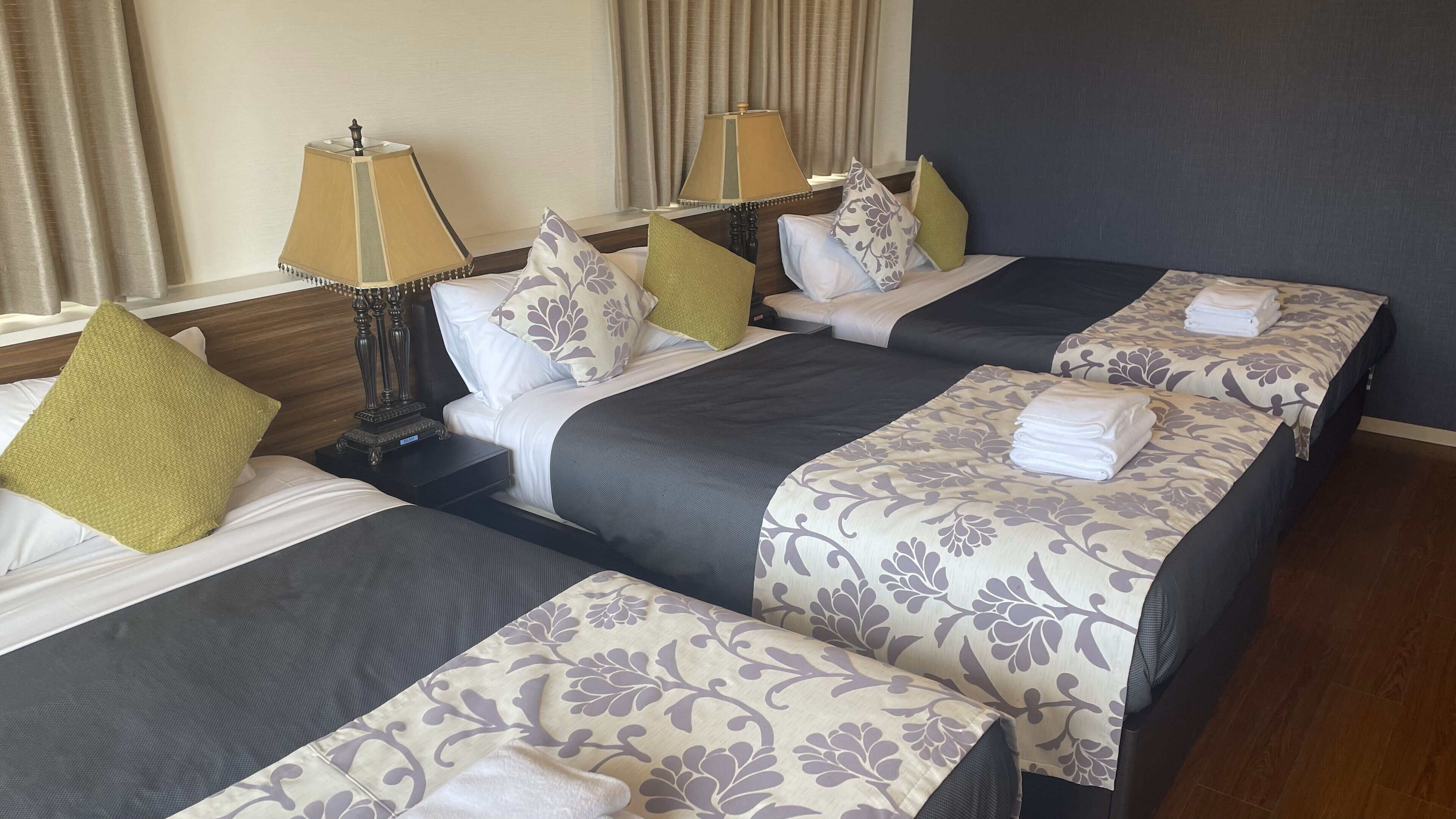 Habitación de hotel con dos camas, cada una con cubrecamas grises oscuros y correpasillos florales. Hay dos lámparas de noche con pantallas, y la habitación tiene un cabecero de madera.