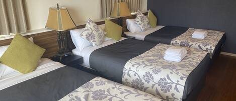 Habitación de hotel con dos camas, cada una con cubrecamas grises oscuros y correpasillos florales. Hay dos lámparas de noche con pantallas, y la habitación tiene un cabecero de madera.