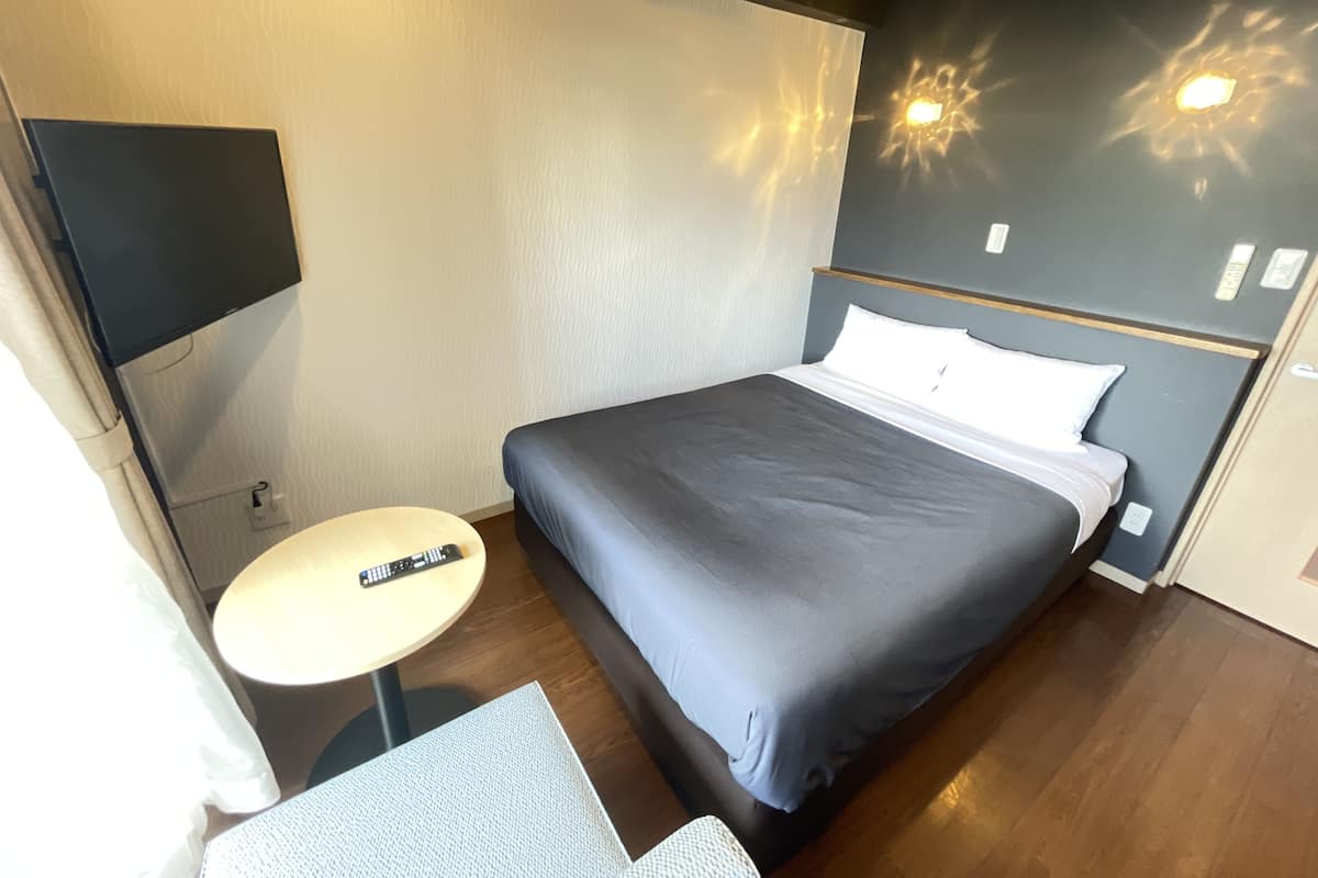 Ein Hotelzimmer mit einem Bett, einem kleinen runden Tisch, einem Sessel, einem an der Wand befestigten Fernseher und einer Wandleuchte.