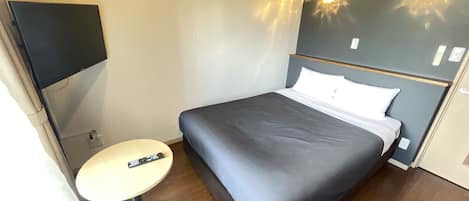 Una habitación de hotel con una cama, una mesita redonda pequeña, una silla, un televisor montado en la pared y una lámpara también fijada a la pared.