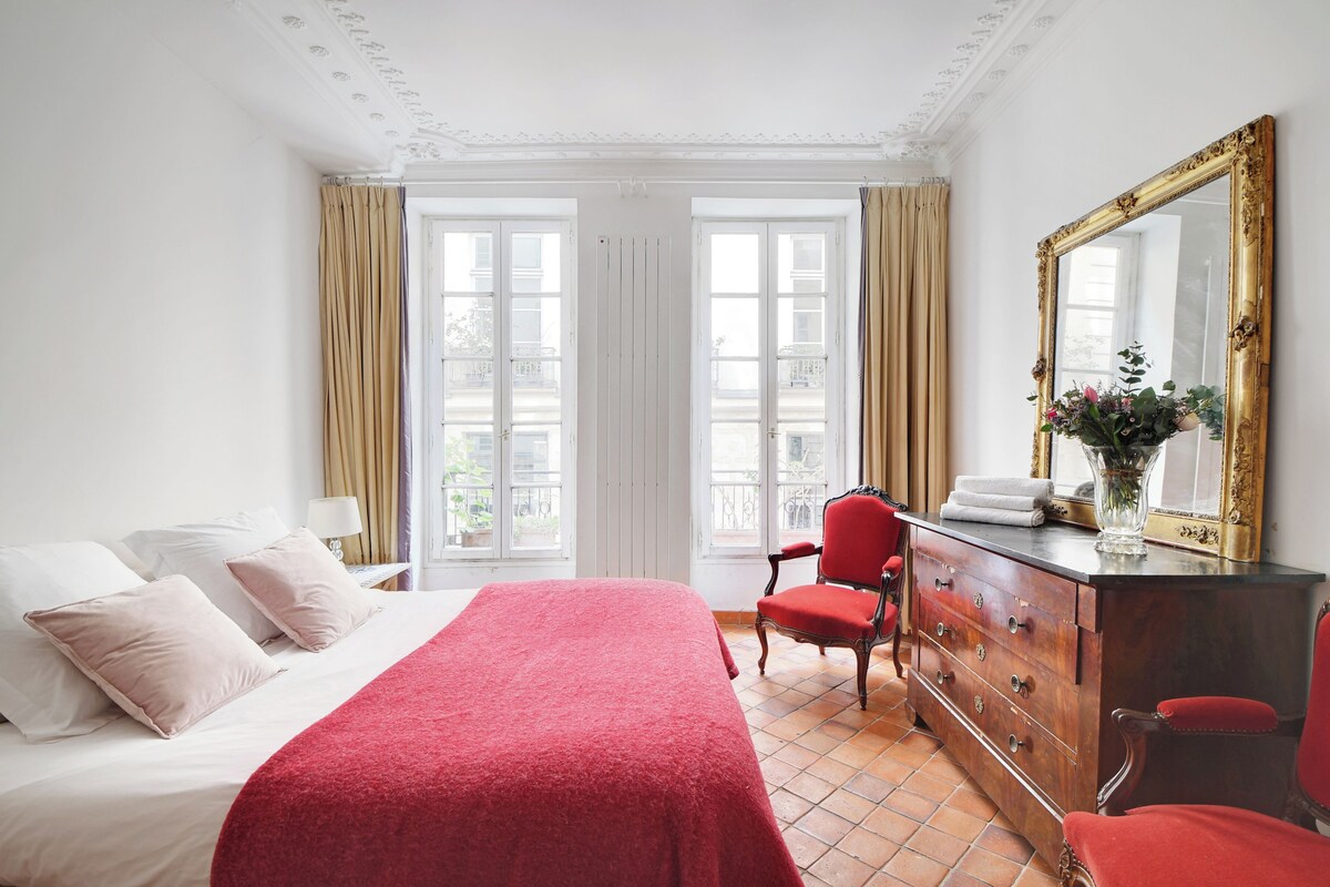 Quarto de hotel com cama, penteadeira, cadeira, espelho e janela com cortinas.