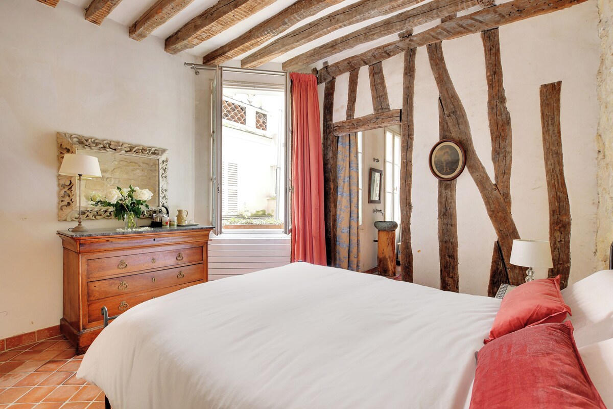 Un dormitorio con techo de madera, cama con ropa de cama blanca, cómoda con espejo y ventana con cortinas.