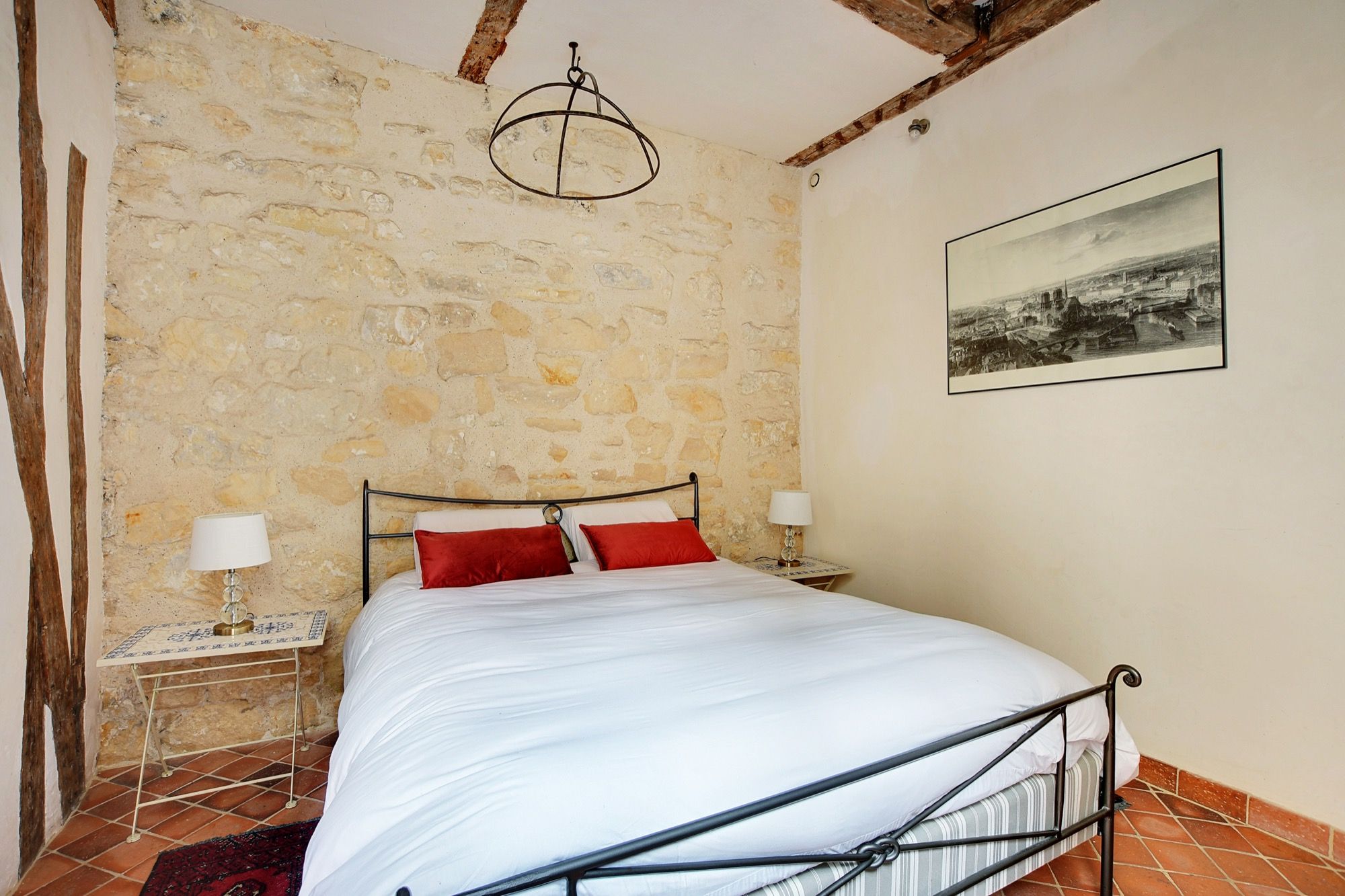A bedroom with a stone wall, a bed with red and white bedding, a hanging light fixture, a nightstand with a lamp, and a framed picture on the wall.