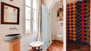 A bathroom with a stone sink, a wooden stool, a shower with a colorful diamond-patterned curtain, and a toilet with a shelf above it.