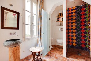 Un baño con un lavamanos de piedra, un taburete de madera, una ducha con una cortina de diseño colorido a rombos y un inodoro con un estante encima.