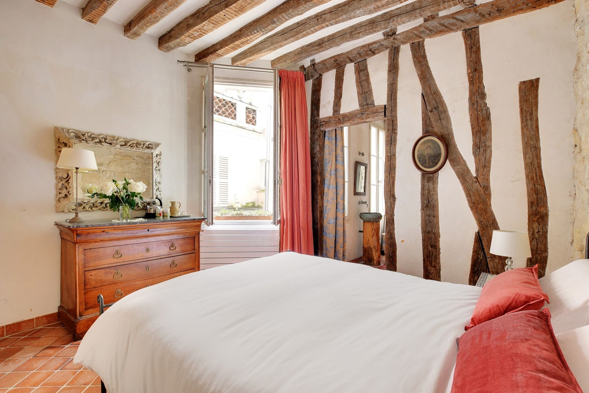 Un dormitorio con vigas de madera a la vista, una cama con ropa de cama blanca, un aparador con cajones, un espejo y una ventana con cortinas.