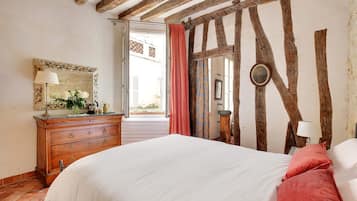 Un dormitorio con vigas de madera a la vista, una cama con ropa de cama blanca, un aparador con cajones, un espejo y una ventana con cortinas.