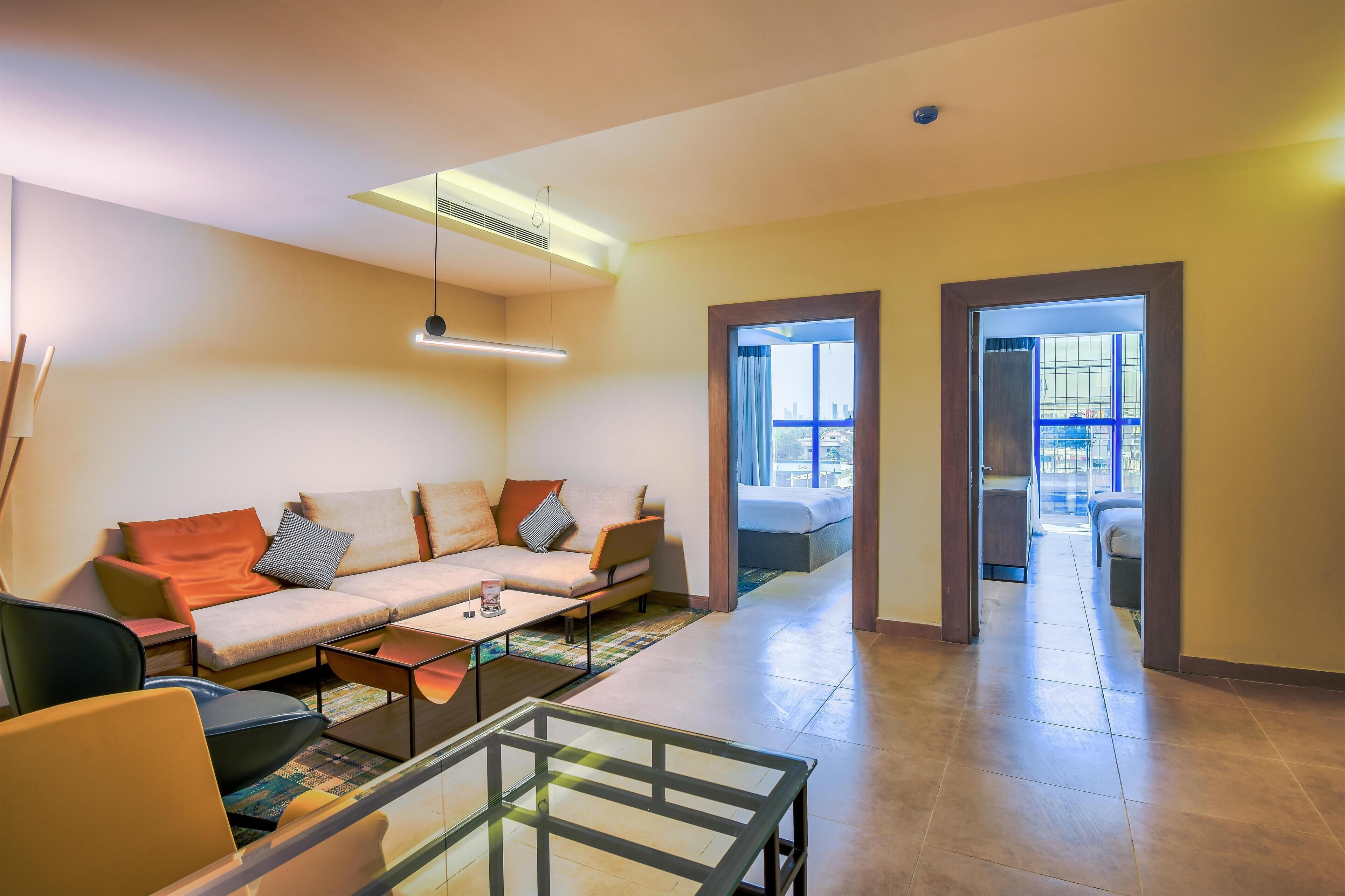 Una sala de estar moderna con un sofá, sillas y una mesa de centro de vidrio. Hay un ventanal con cortinas y vistas a la ciudad.