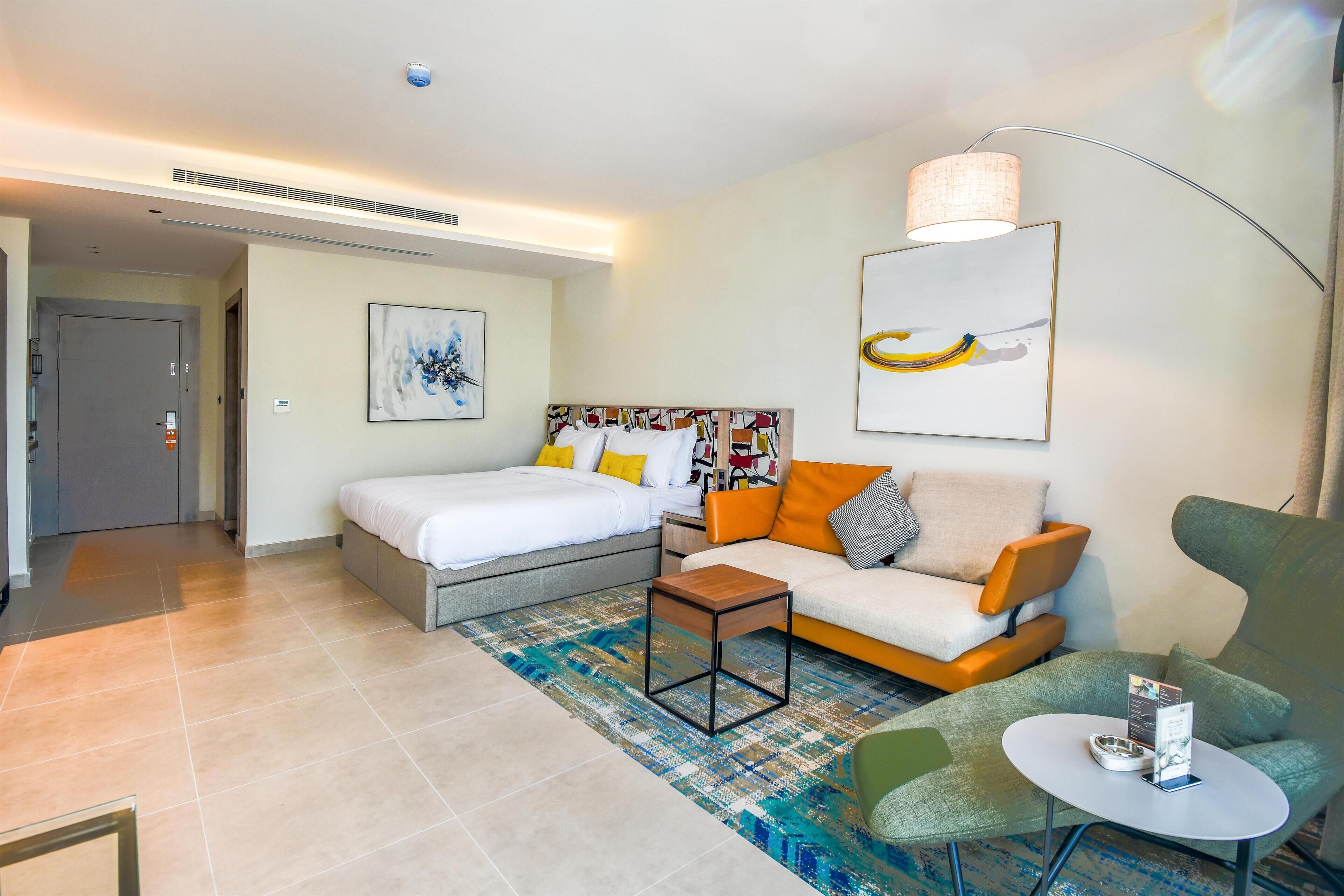 A modern hotel room with a bed, a sofa, a small table, and a chair. There is a large window and a door leading to the hallway.