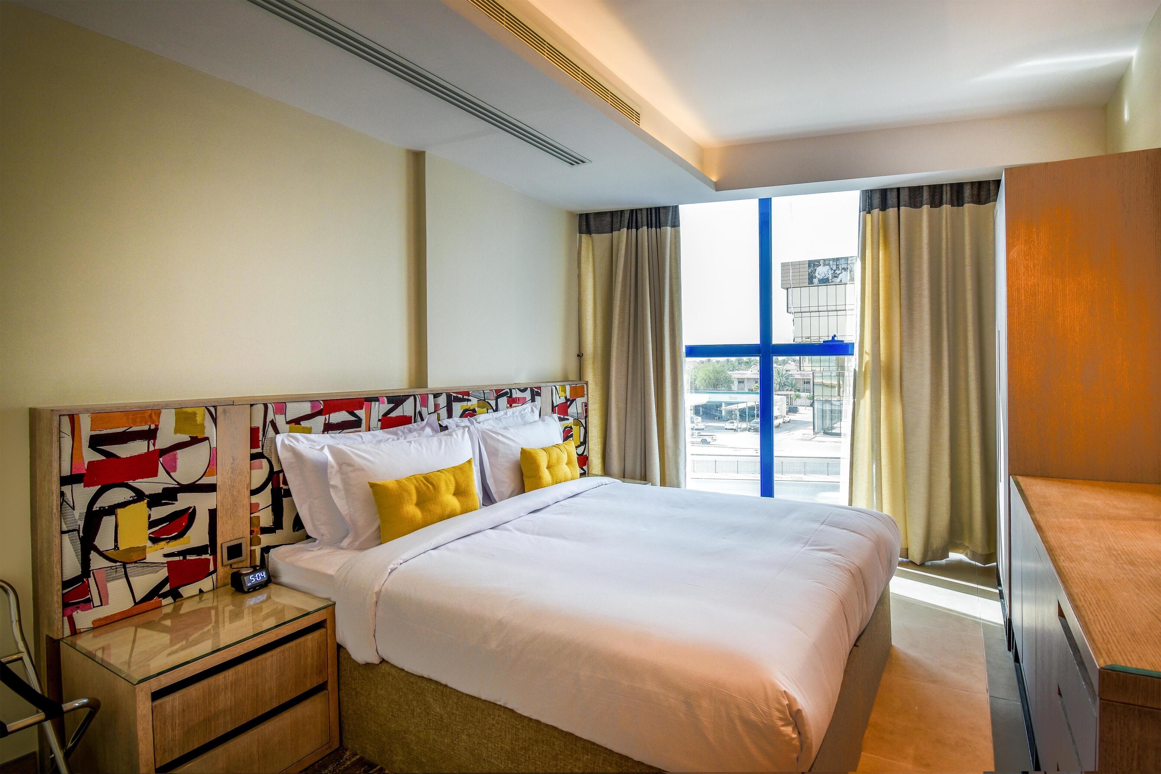 A hotel room with a large bed, a bedside table with a clock, a modern abstract wall art, and a view of the city through large windows.