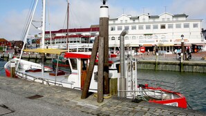 A waterfront hotel with a docked boat, a lighthouse, and a row of shops in the background. - Apartment Strandstrasse 10 / Büsum (Büsum)