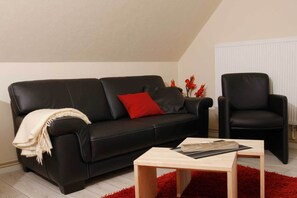En svart lädersoffa med en röd kudde, ett träbord och en röd matta i ett rum med ett sluttande tak och ett element.
