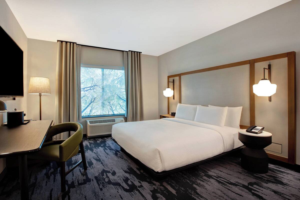 A hotel room with a bed, a desk with a chair, a TV, a lamp, and a window with curtains.