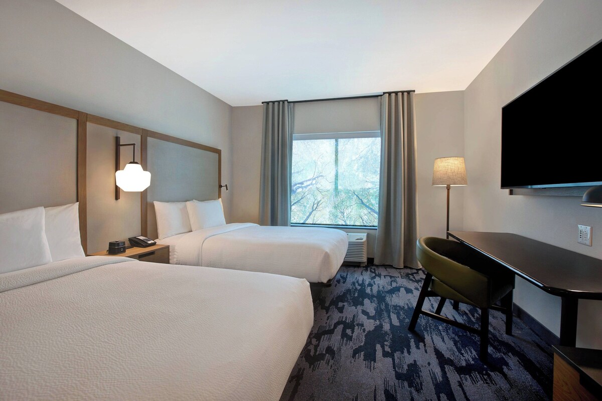 A hotel room with two beds, a desk, a flat-screen TV, and a window with curtains.