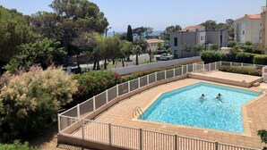 Una piscina con dos personas nadando, rodeada por una cerca blanca, con edificios y árboles al fondo.