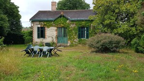 Une maison de campagne au cachet rustique, avec une façade en pierre et des volets verts, entourée d’une végétation luxuriante et d’une pelouse verdoyante où se trouvent une table et des chaises pour les repas.