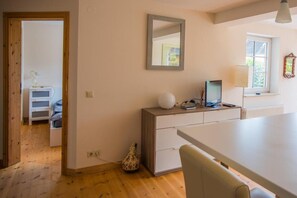 Ein modernes Zimmer mit Holzboden, einem weißen Schreibtisch, einem Stuhl, einer kleinen Kommode, einem Spiegel und einem Fenster mit Jalousien.
