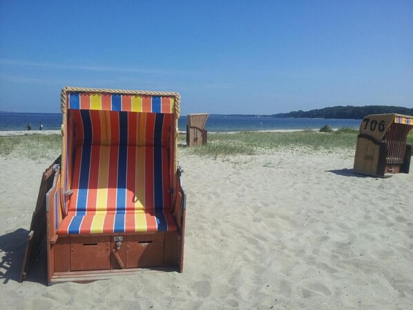 Ein Liegestuhl mit gestreifter Markise, ein Holzstuhl und ein Schild mit der Nummer 106 an einem Sandstrand.