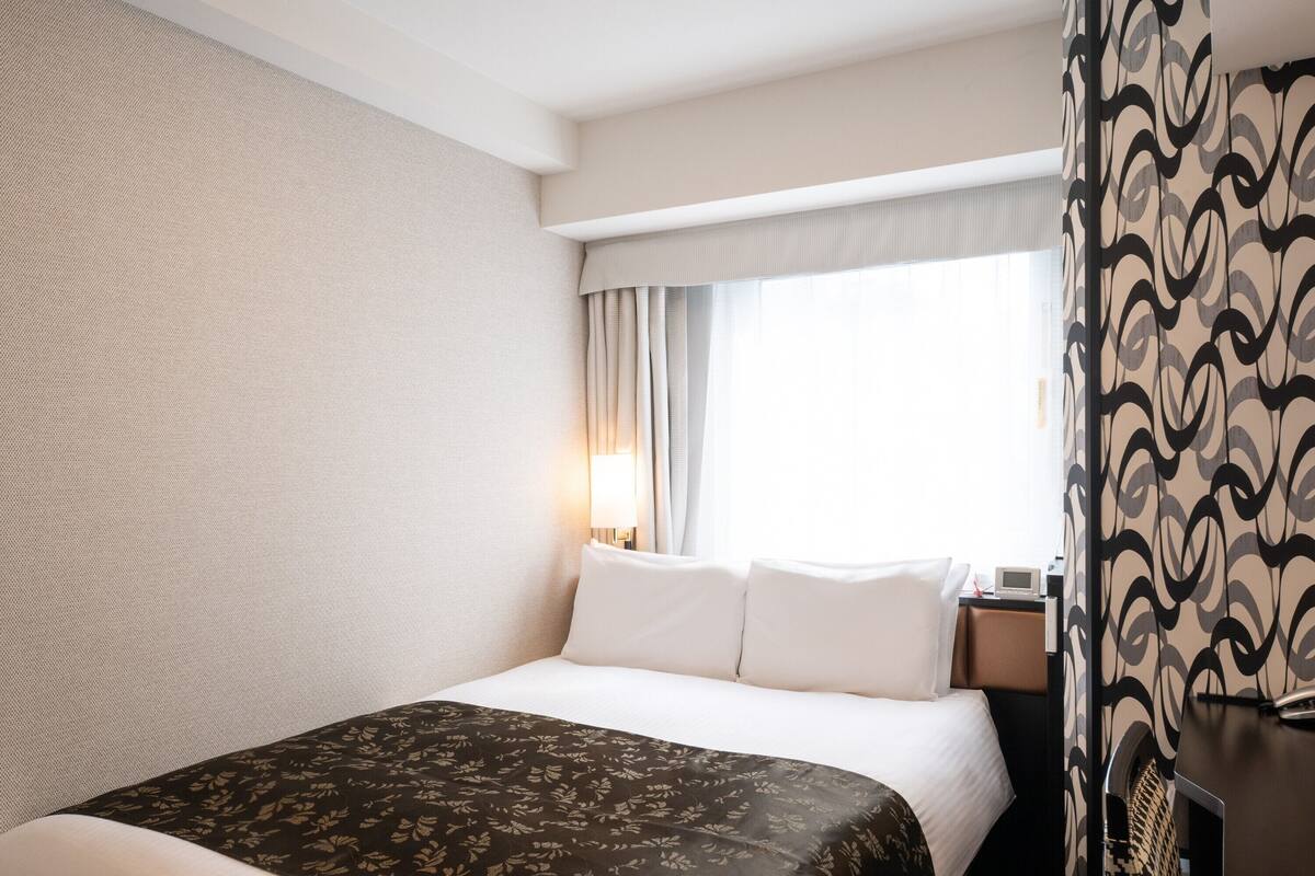 A hotel room with a bed, a nightstand, a lamp, and a window with curtains.