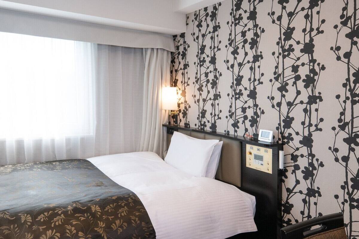 A hotel room with a bed, a nightstand with a lamp, and a wall with a floral pattern.
