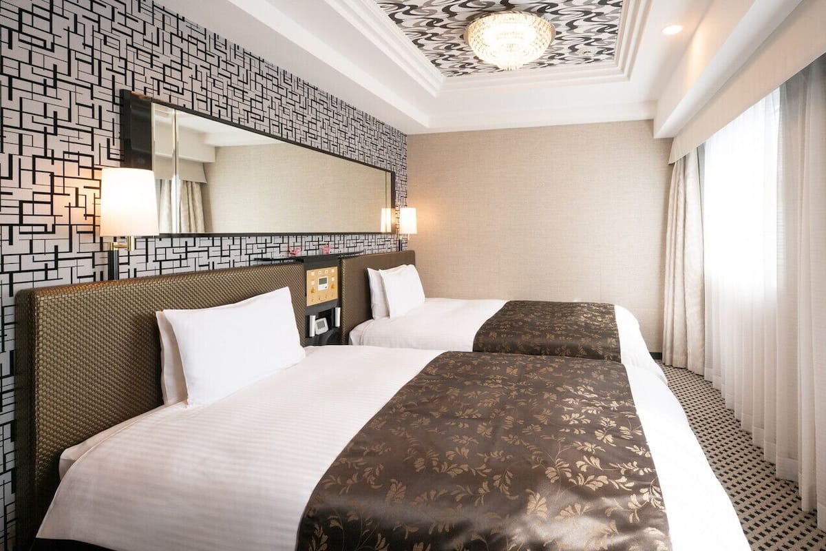 A hotel room with two beds, a large mirror, a patterned wall, and a window with curtains.