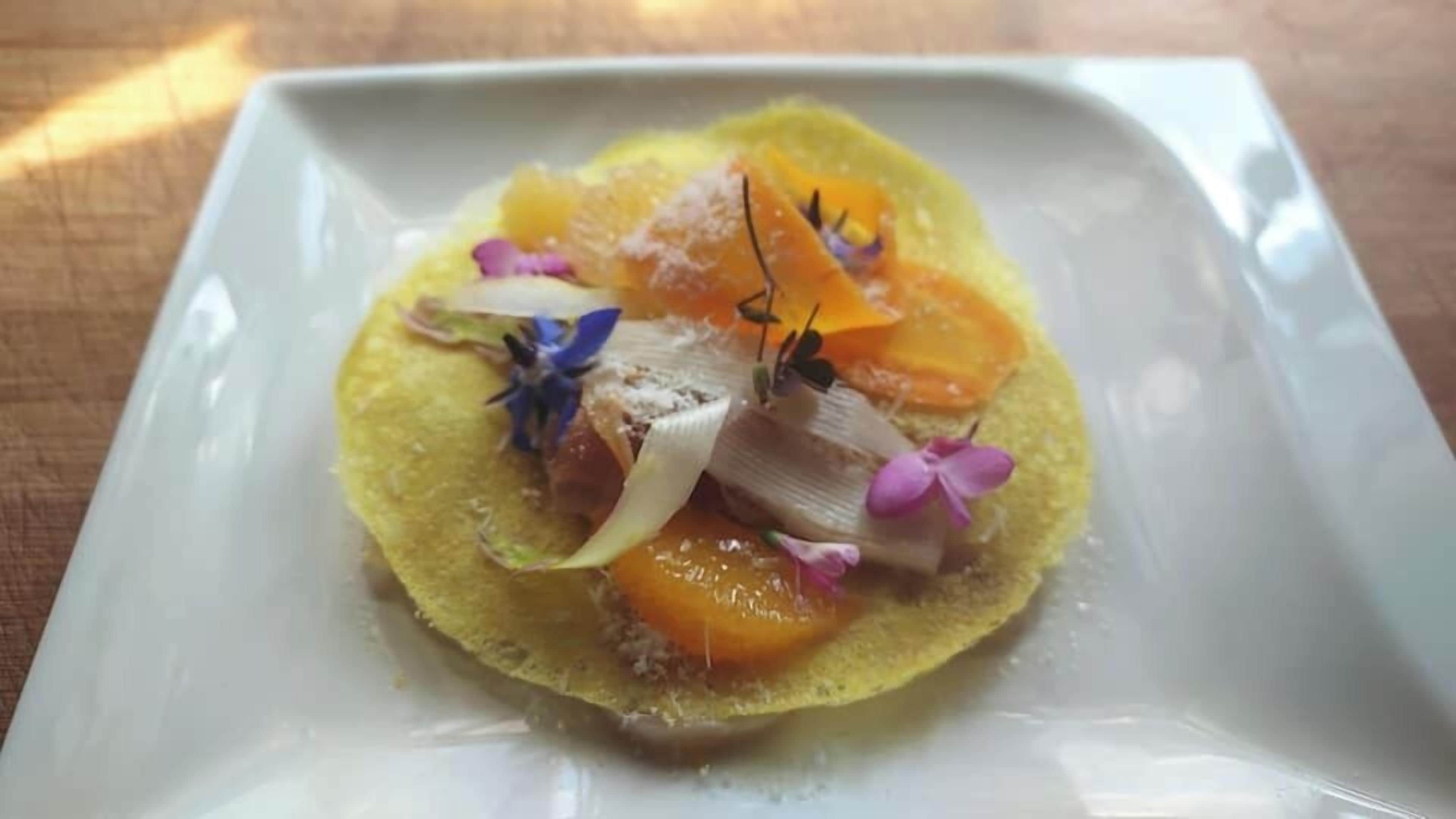 Un plato cuadrado blanco con base amarilla, cubierto de rodajas de naranja, cebolla blanca, flores púrpuras y un ramito de verdura.