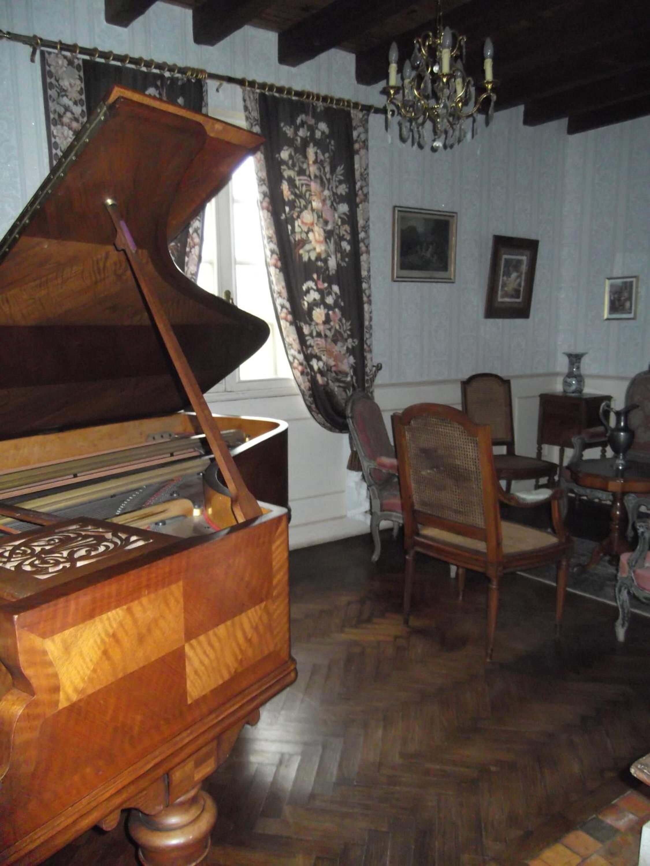 Un piano à queue se trouve dans une pièce dotée d’un plancher en bois, d’un lustre, de rideaux à motifs floraux et d’un coin repas avec des chaises et une table.