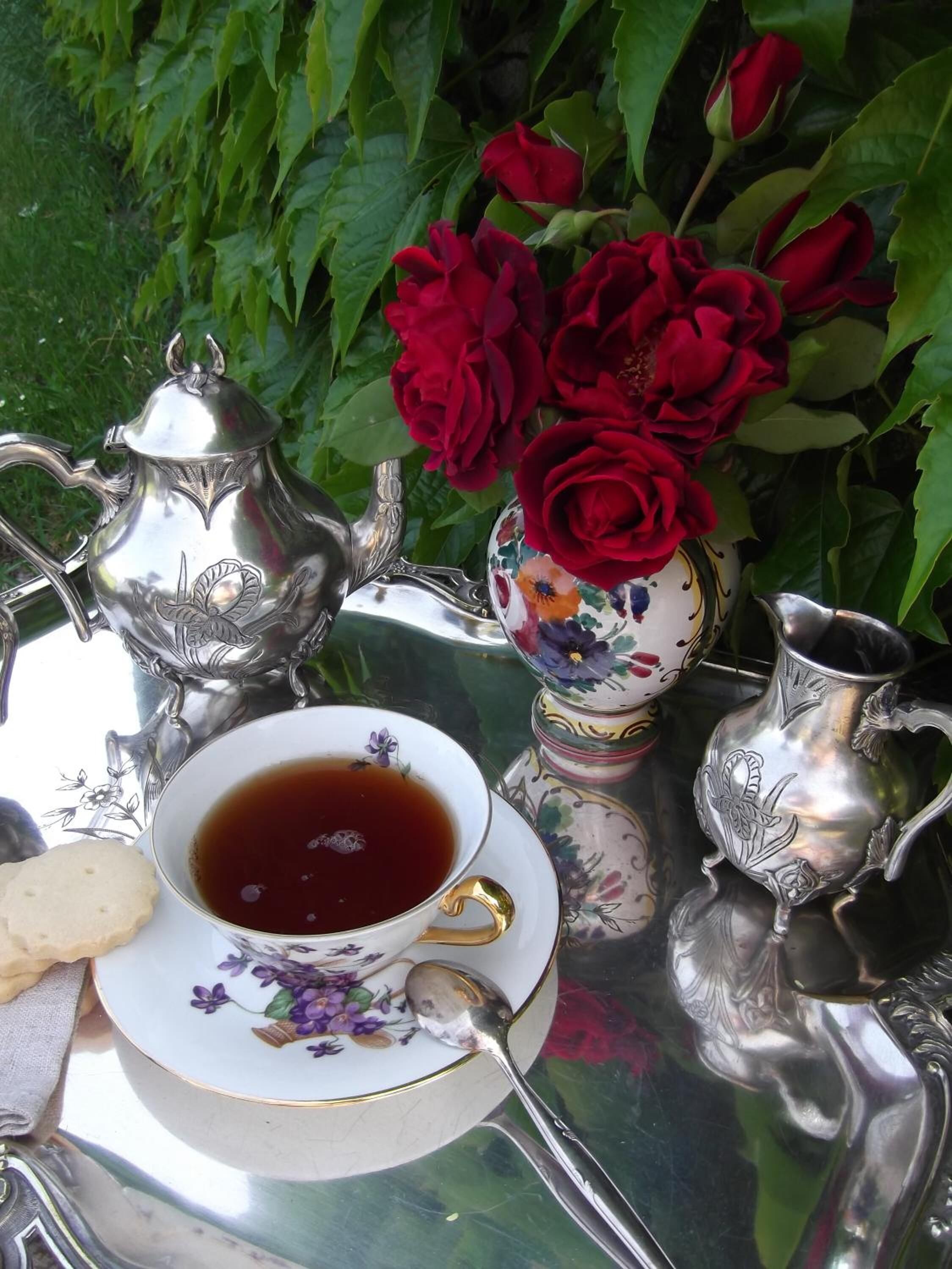 A table set with a teapot, cup, saucer, and sugar bowl, surrounded by red roses and a decorative vase.