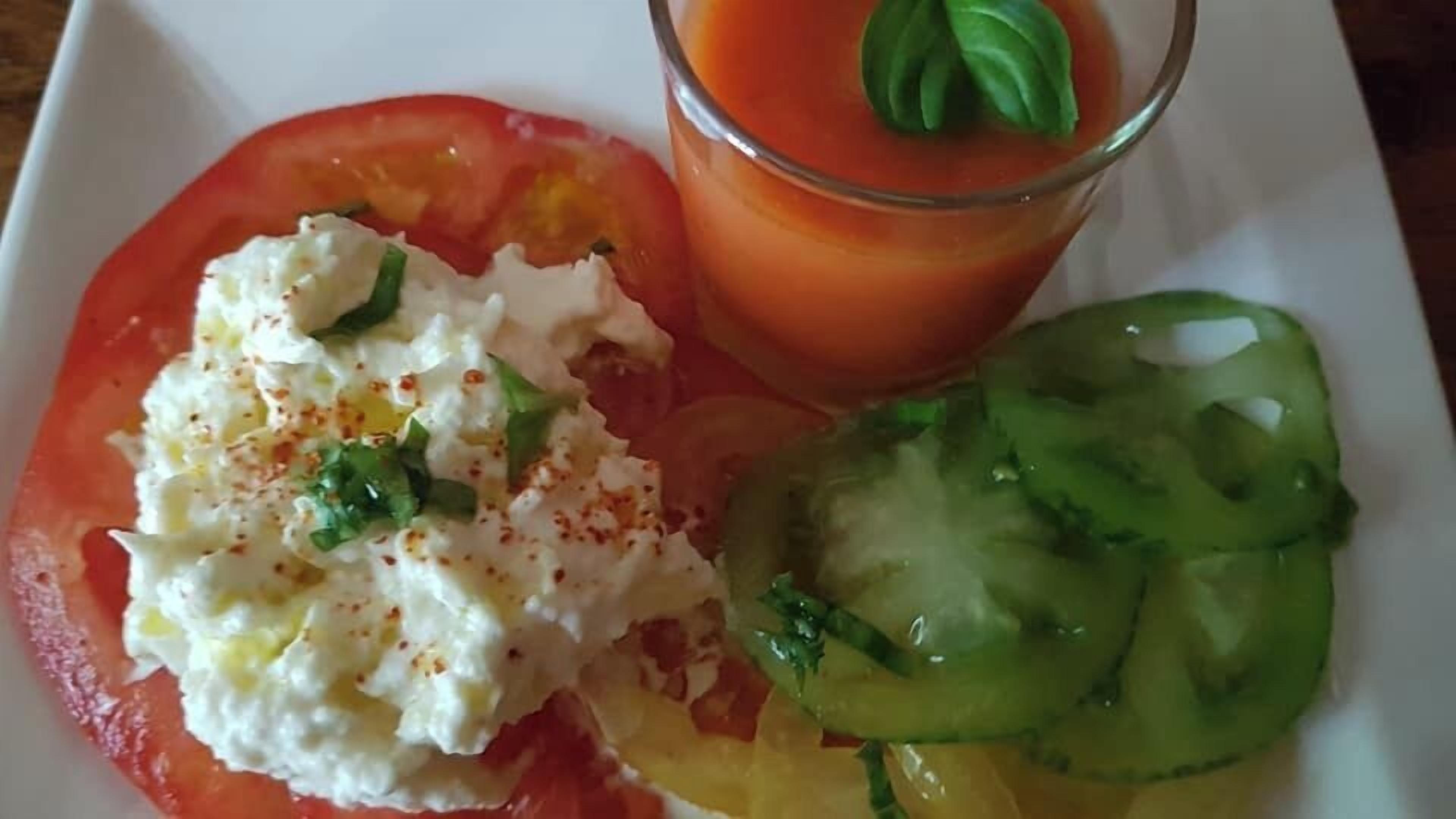 Une assiette avec une tranche de tomate recouverte d’une substance blanche et crémeuse, un verre de jus de tomate orné d’une feuille de basilic et une tranche de concombre.