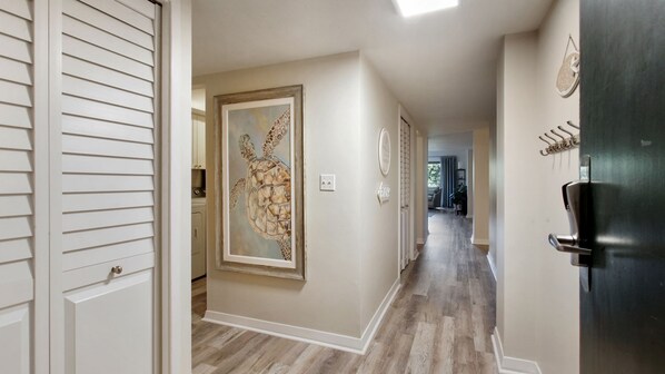 A hallway with a wooden floor, a framed artwork on the wall, a door with a handle, and a clock on the wall.
