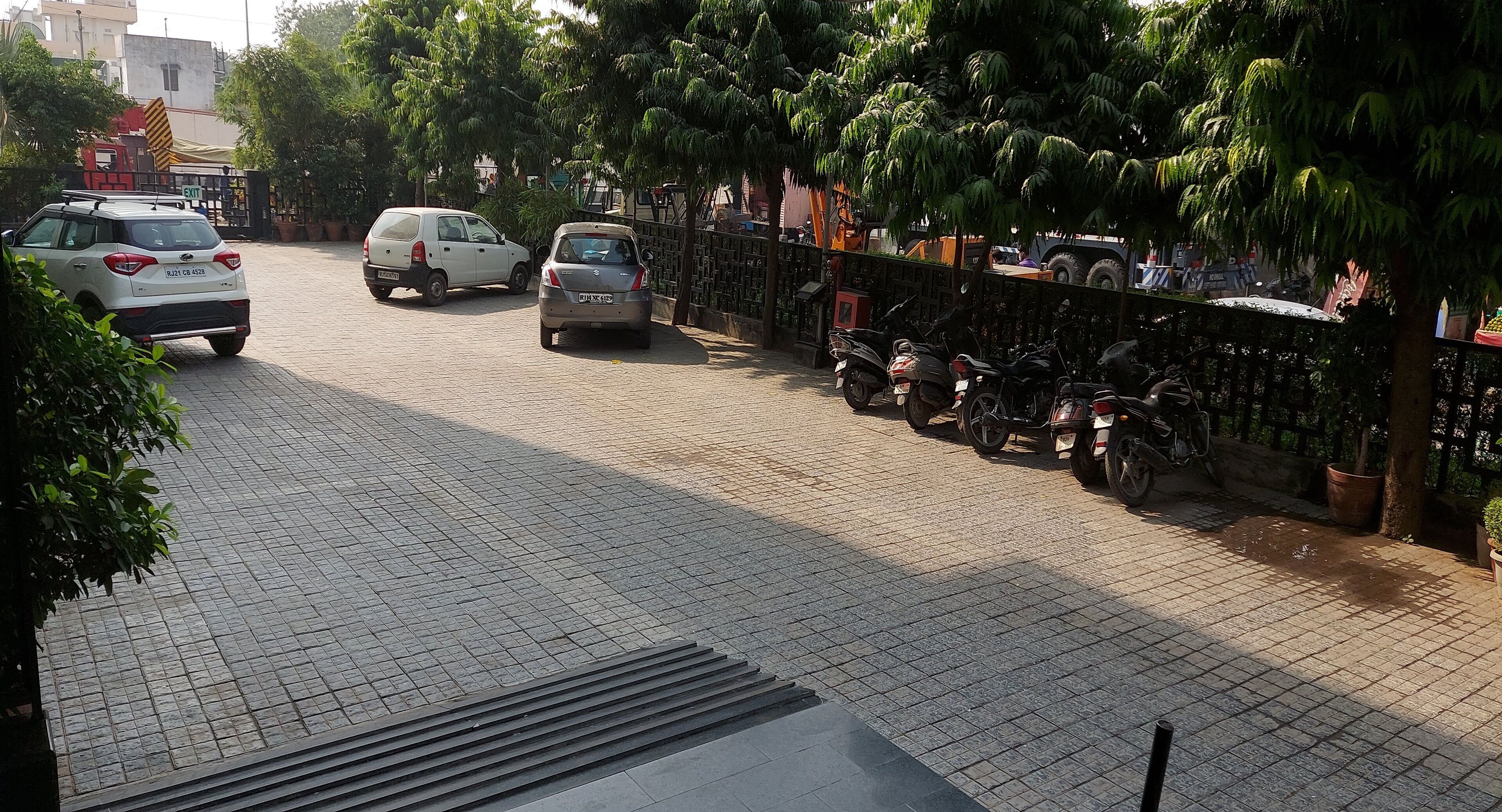 A paved street with parked cars and motorcycles, trees, and a fence.