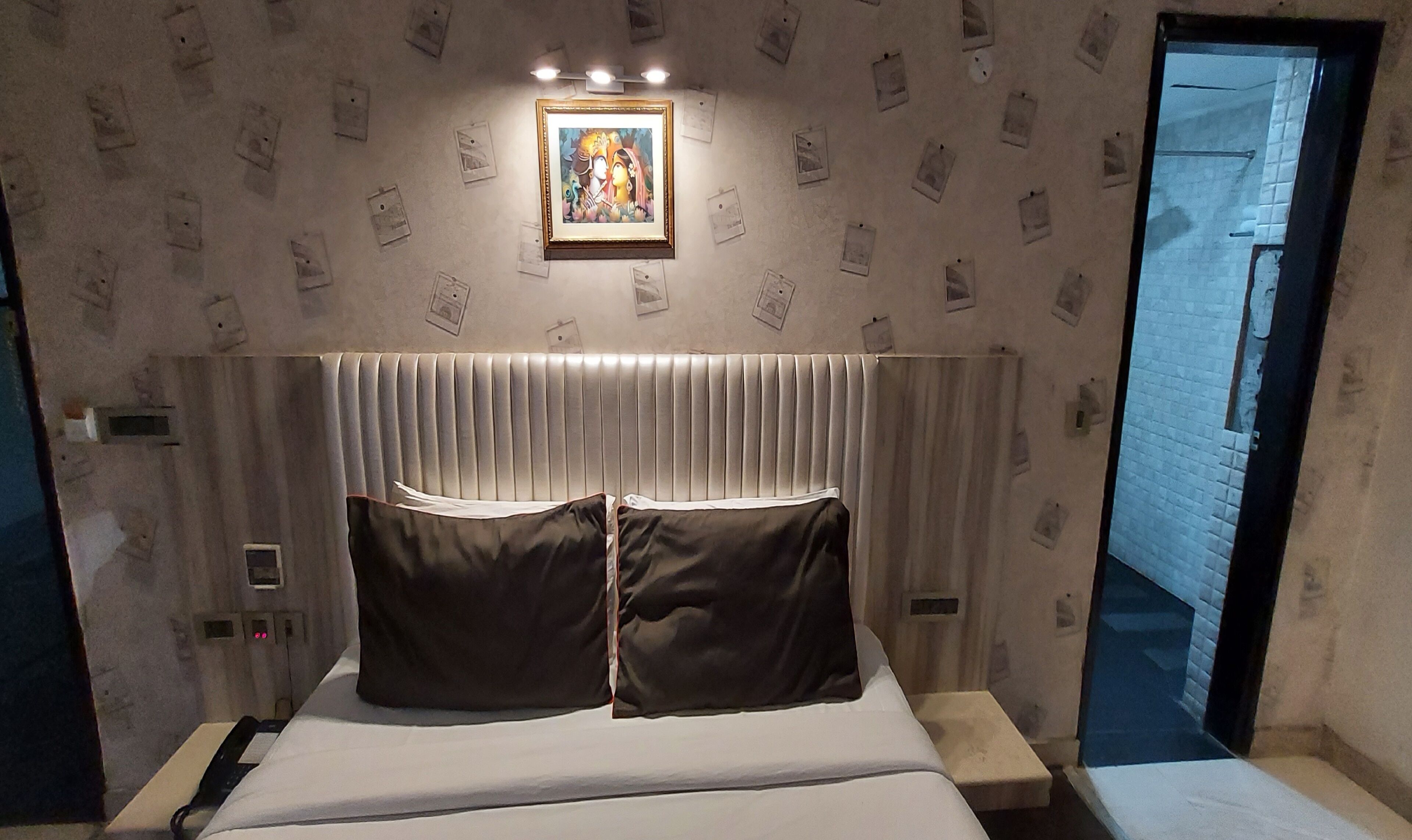 A hotel room with a bed, a headboard with vertical slats, a bedside table, a nightstand, a framed picture on the wall, and a door leading to a corridor.