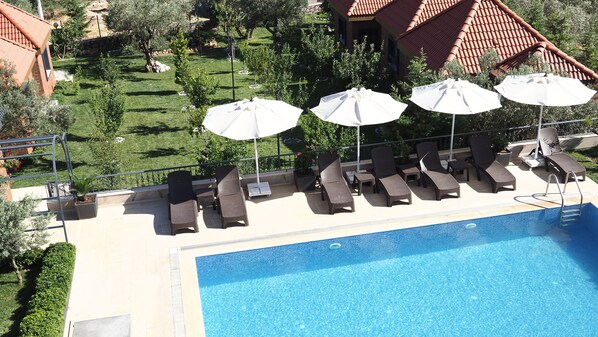A poolside area with lounge chairs and umbrellas, a clear blue pool, and a garden with trees and a lawn.