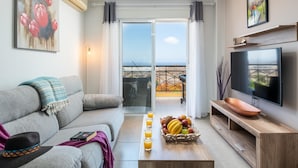 Uma sala de estar moderna com um sofá cinzento, uma mesa de centro de madeira com frutas, uma televisão de tela plana e uma varanda com vista para o mar.