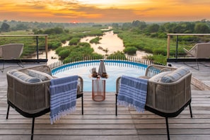 Een houten terras met twee rieten stoelen en een tafeltje met een karaf en beker, met uitzicht op een rivier en de zonsondergang.