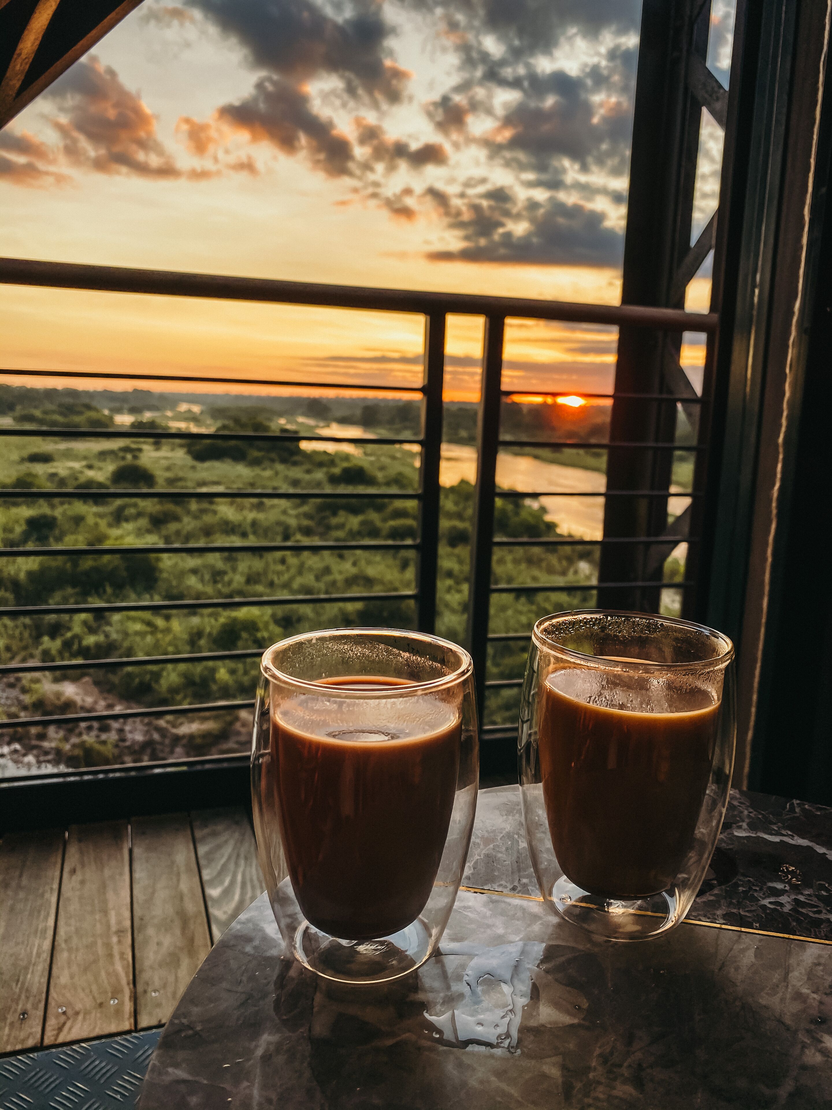 Two glasses of iced coffee on a table with a view of a sunset over a landscape.