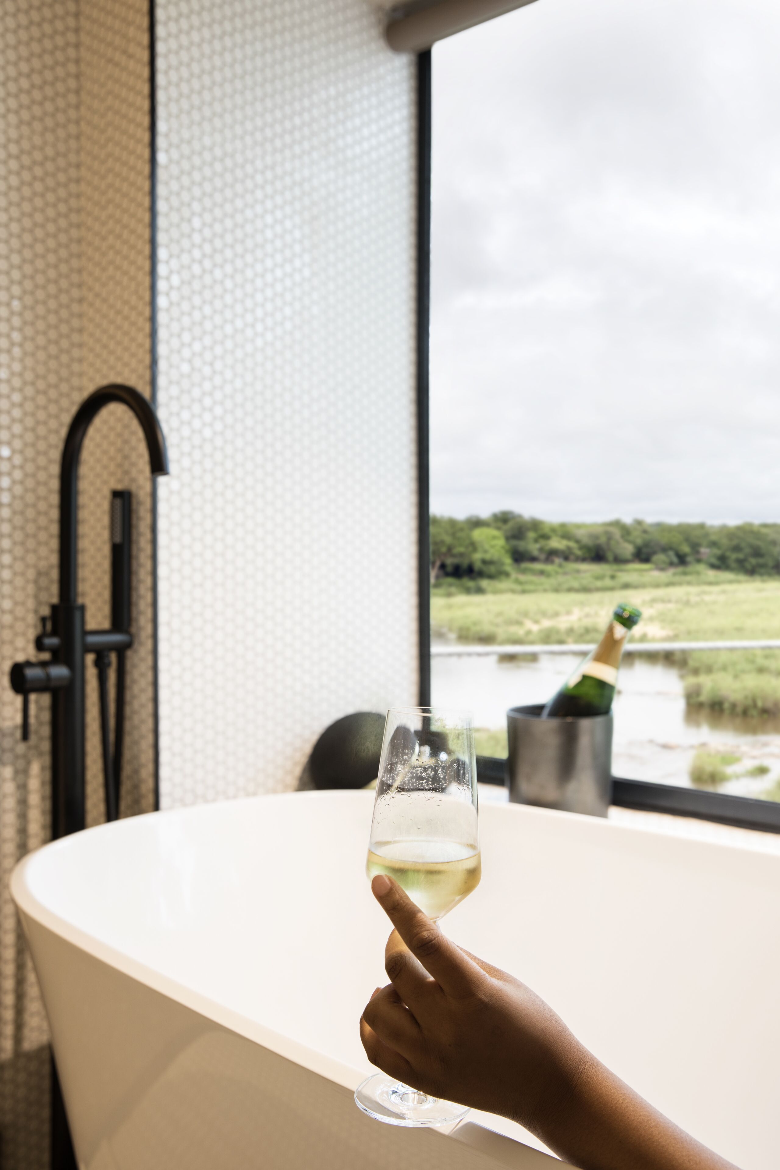 Une main tenant un verre de vin blanc dans une baignoire, avec vue sur une rivière et la verdure environnante.