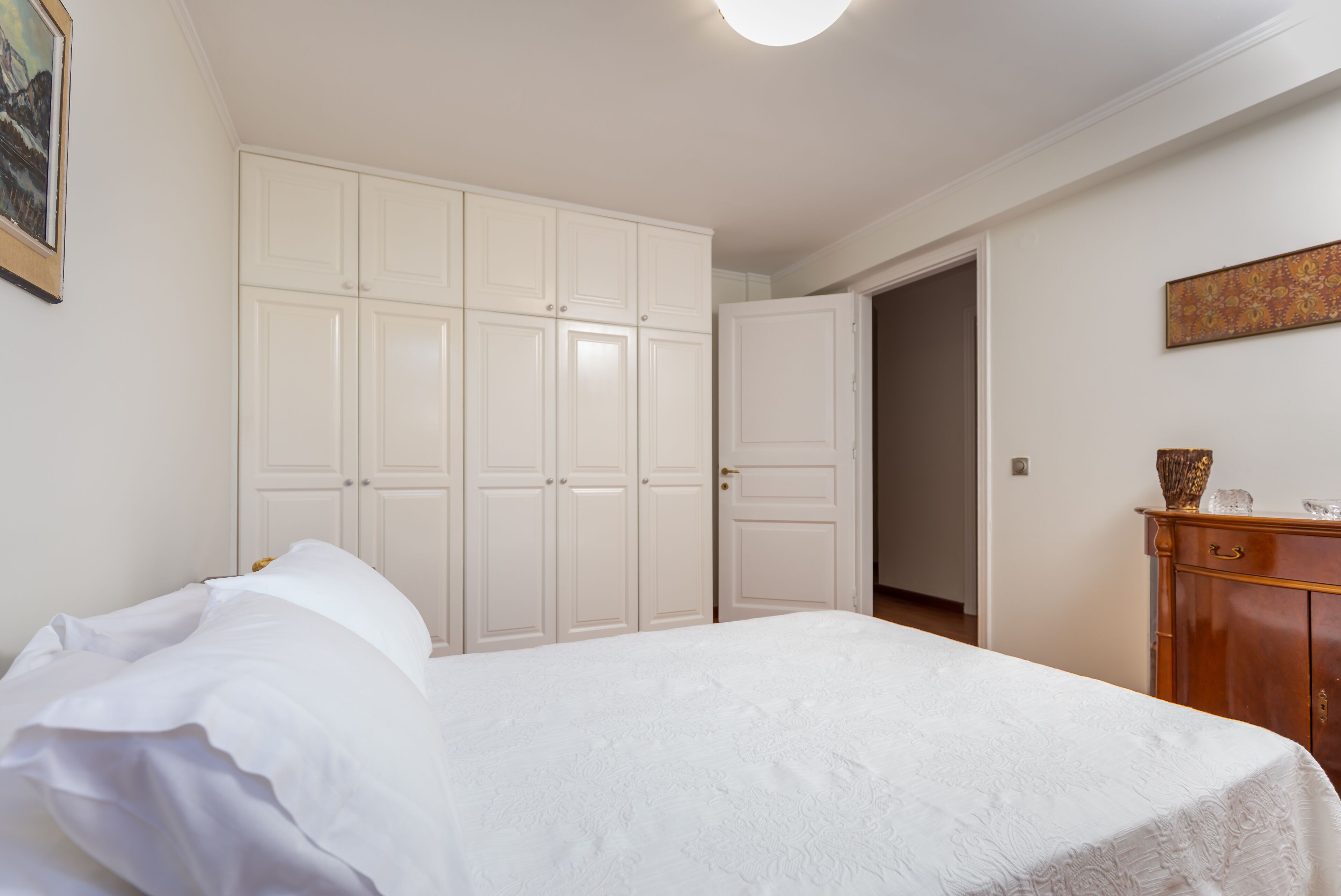 Quarto com cama, guarda-roupa branco e cómoda de madeira.