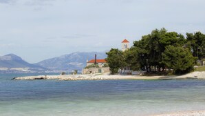 A coastal scene with a stone building and a red-roofed tower, surrounded by trees and a clear view of the sea and distant mountains. - Villa Sv Kriz (Trogir)