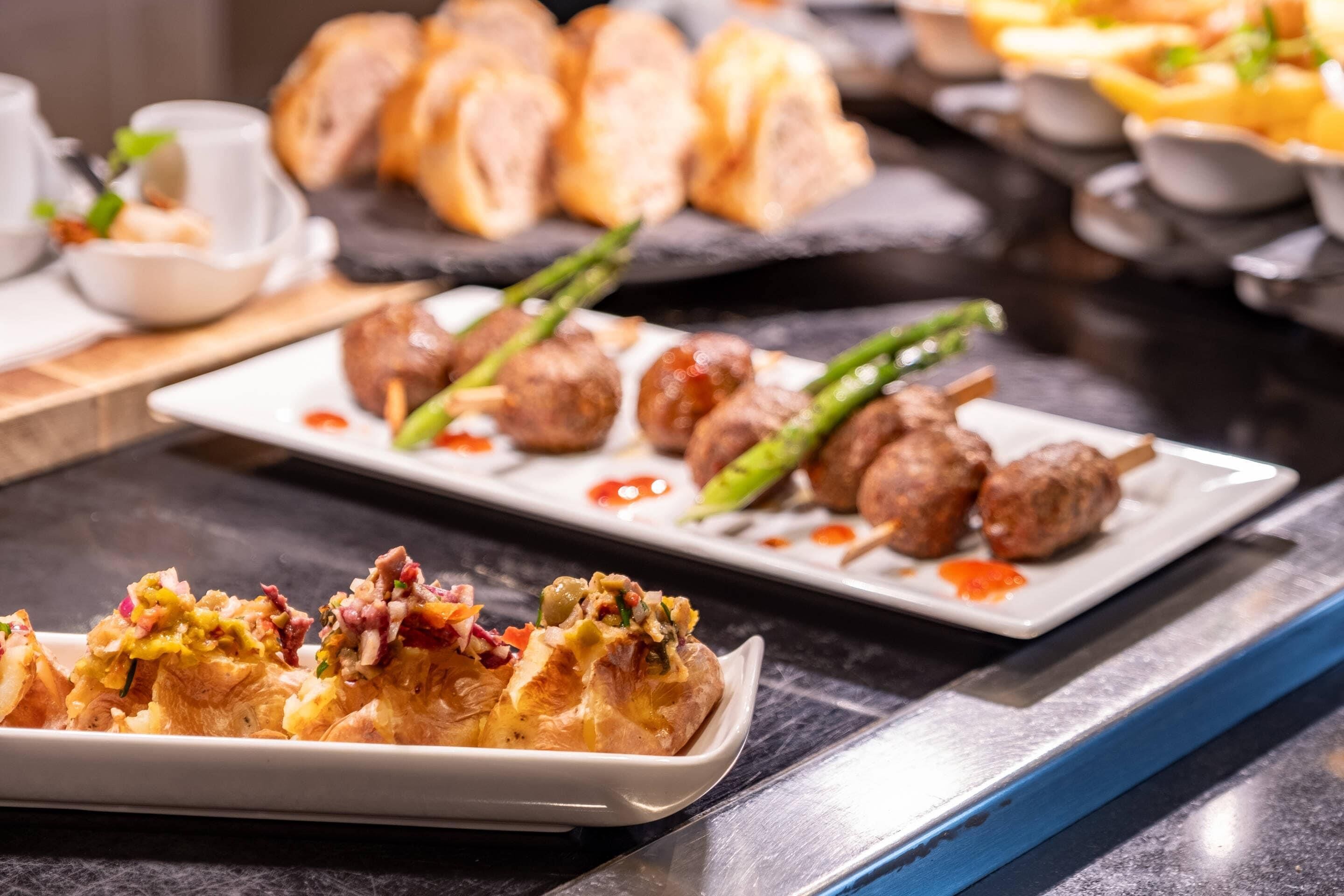 Verschiedene Gerichte auf einem Buffet, darunter spießes Fleisch, eine Auflaufform mit Gemüse und weitere angerichtete Speisen.