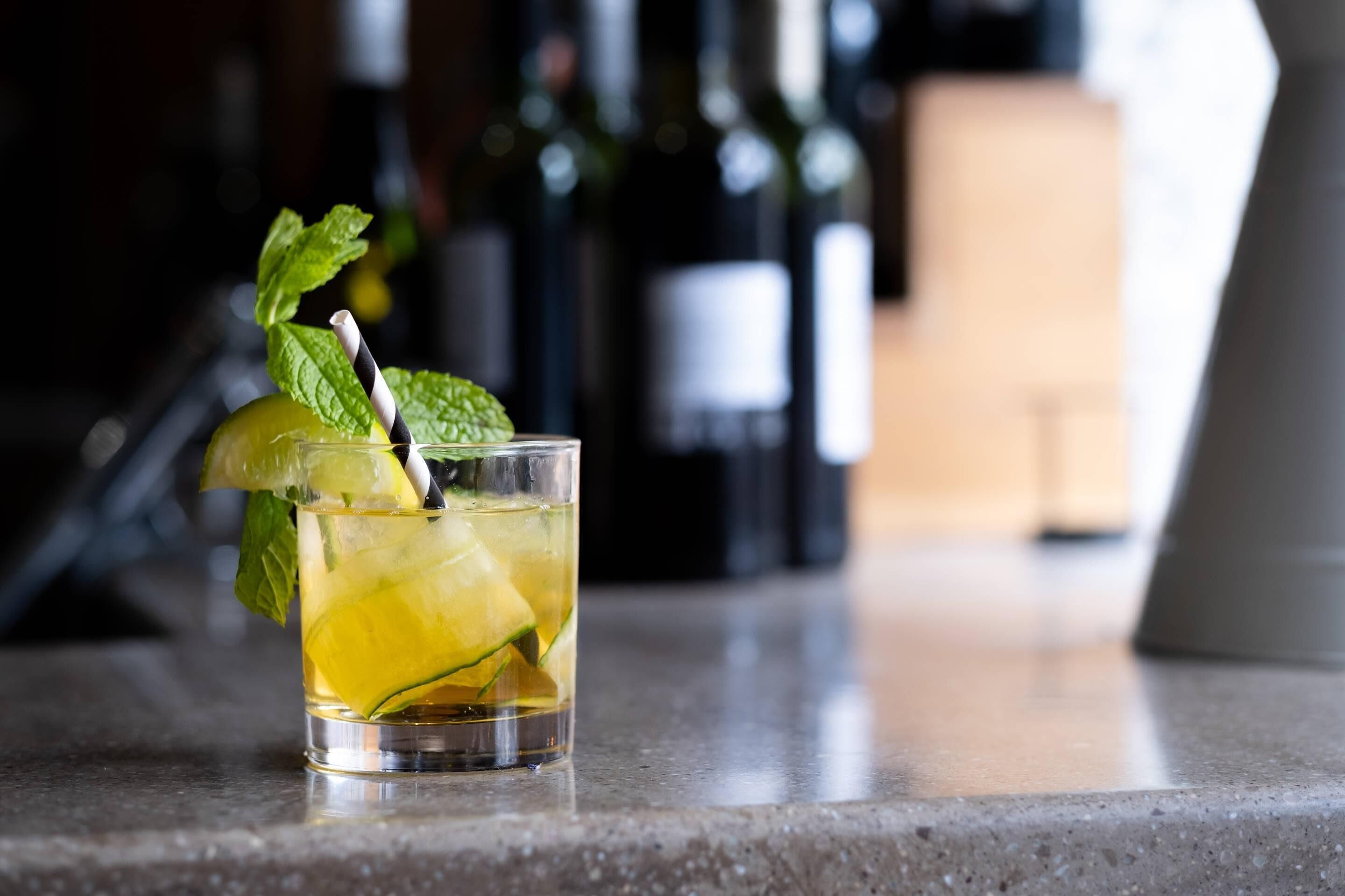 Un vaso de bebida amarilla con hojas de menta y una pajita, sobre una encimera con botellas desenfocadas al fondo.