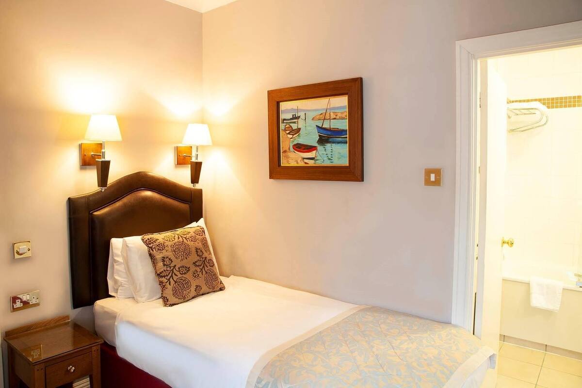 Quarto de hotel com uma cama, duas luminárias na parede, um quadro e uma porta para o banheiro.