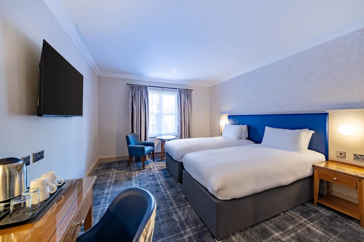 Quarto de hotel com duas camas, uma televisão de tela plana, uma mesa com chaleira e canecas, uma cadeira e uma mesinha.