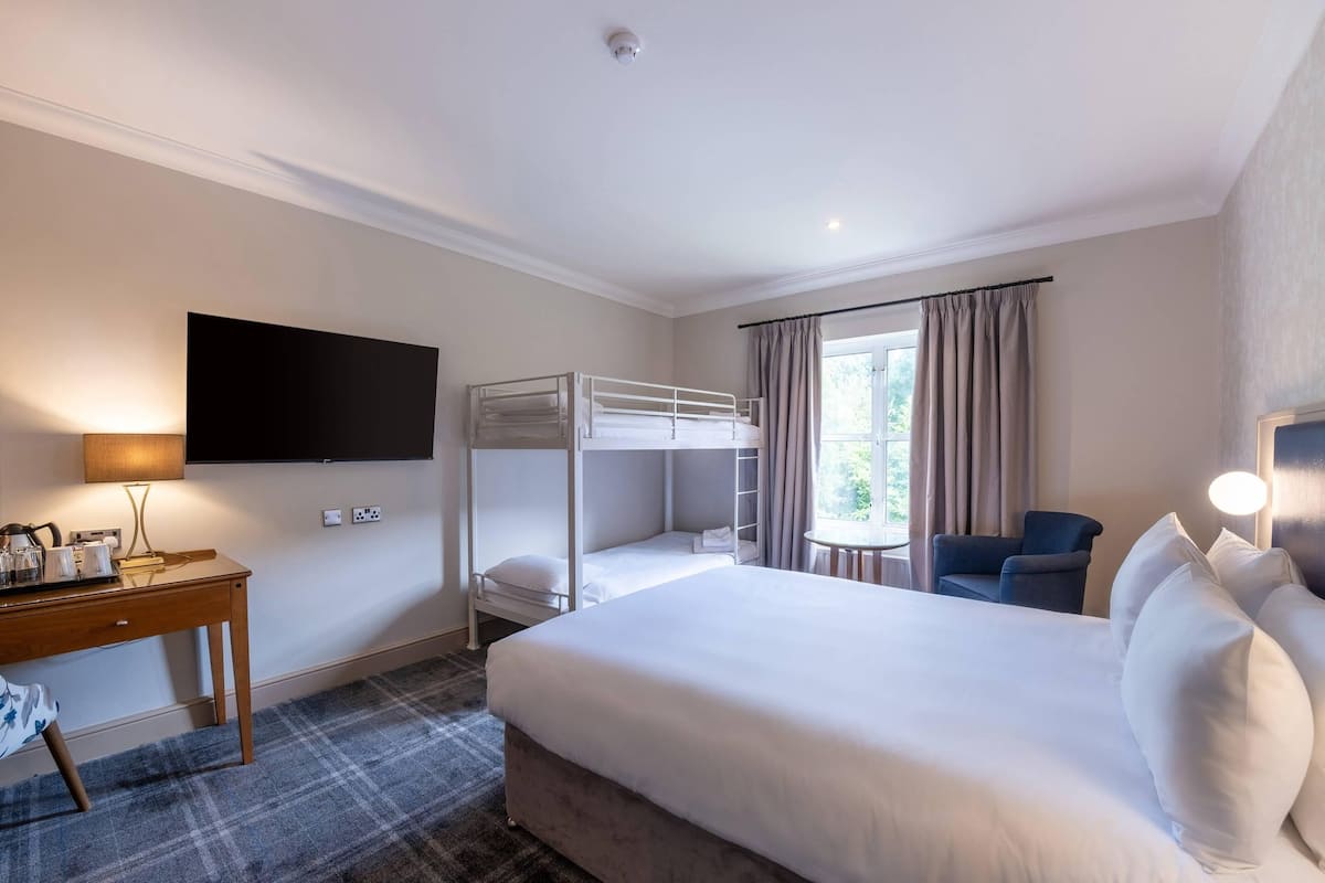 Quarto de hotel com uma cama grande, uma escrivaninha com luminária, uma cama beliche, uma cadeira e uma televisão na parede.
