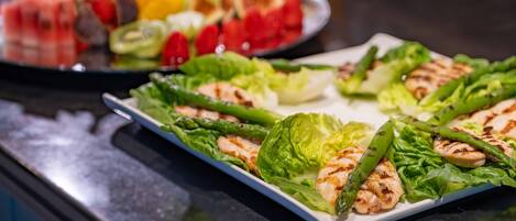 Una mesa de buffet con varios platos, incluyendo pollo a la parrilla, espárragos y frutas variadas.
