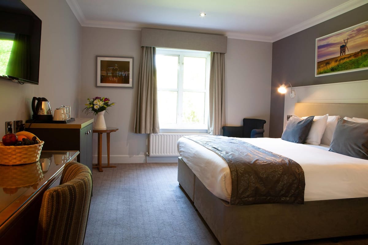 Quarto de hotel com cama, escrivaninha com cesta de frutas, vaso com flores, janela com cortinas e um quadro na parede.