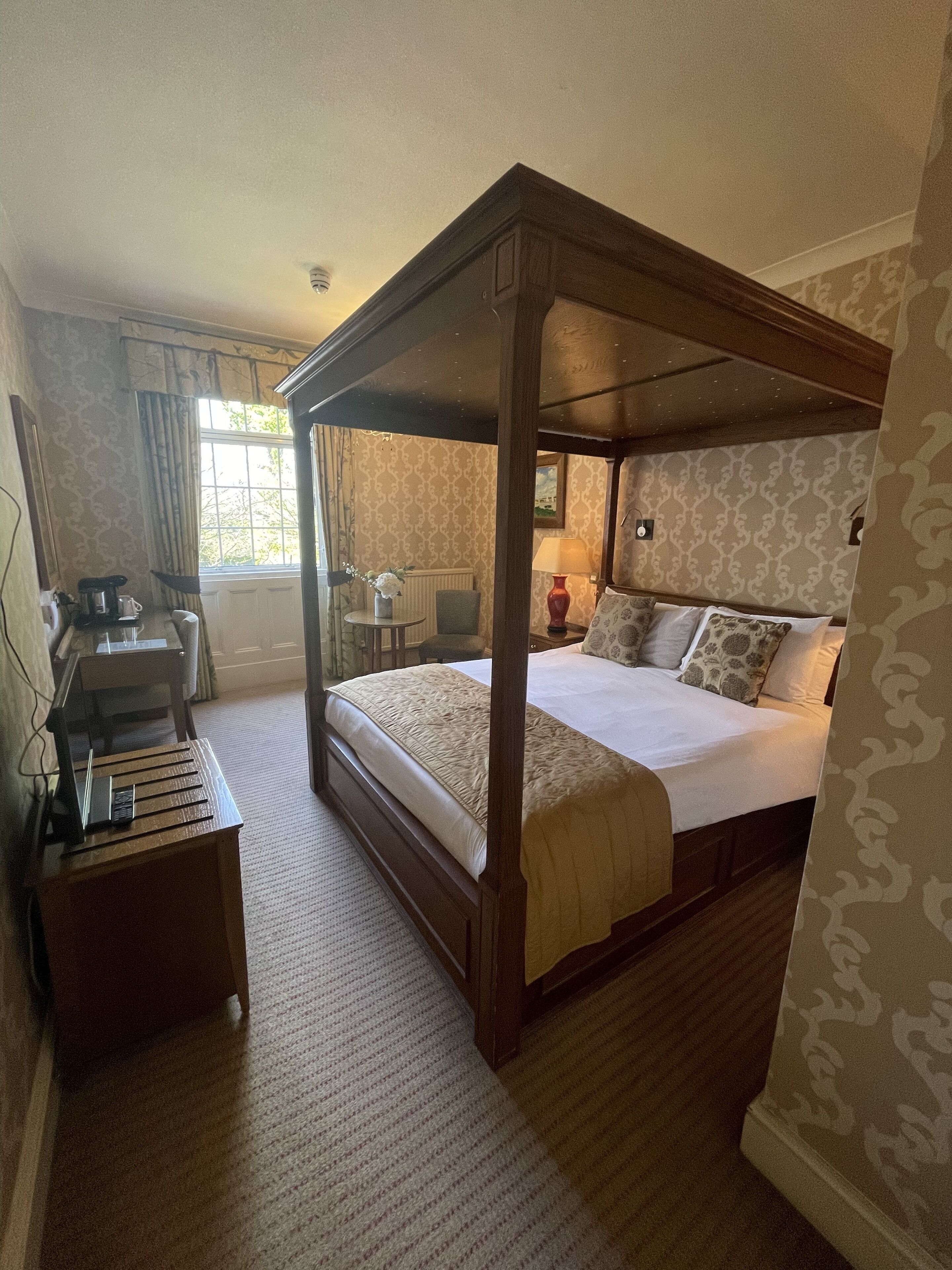Uma cama com dossel e estrutura de madeira, uma mesa de cabeceira com abajur e vista para o exterior pela janela.