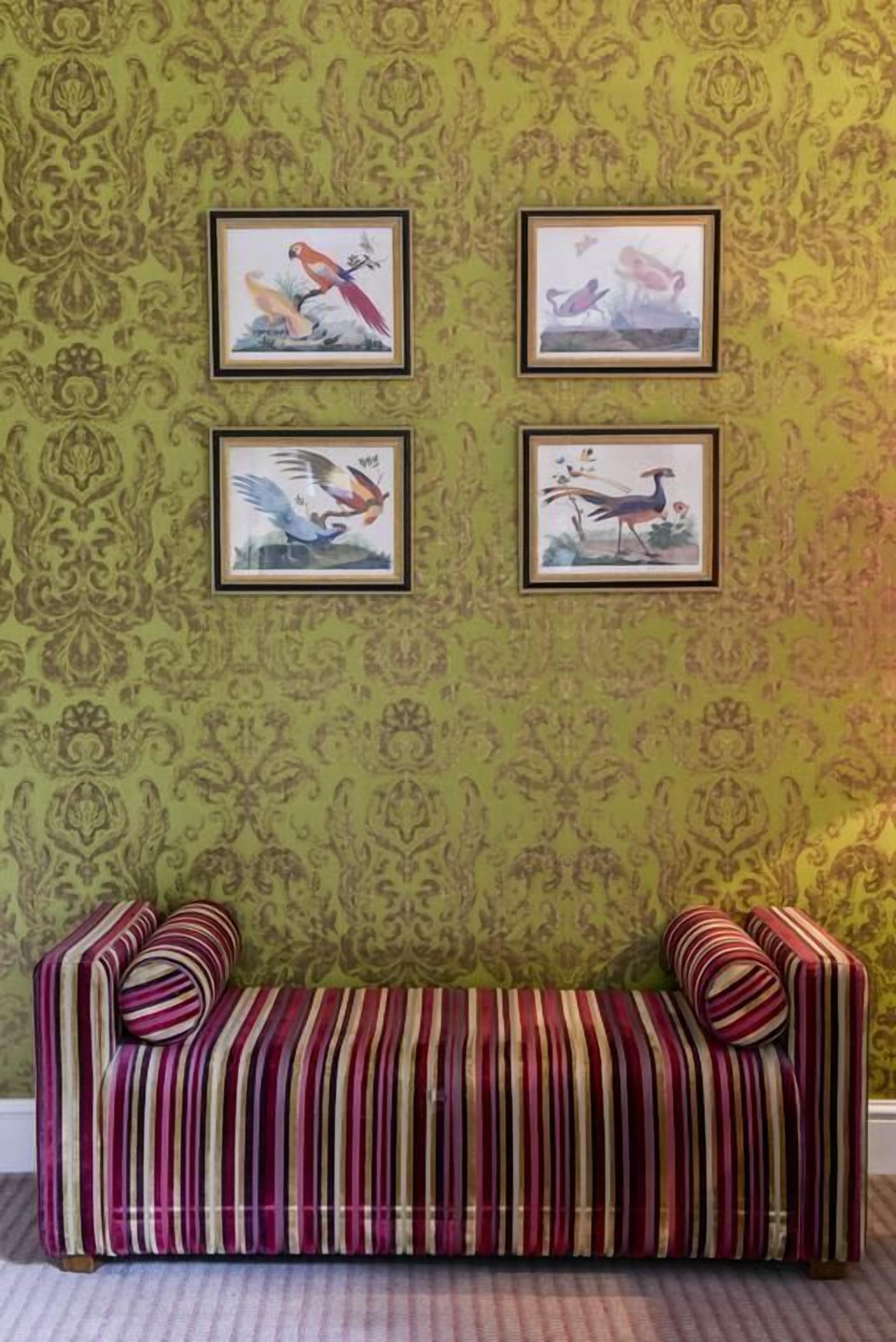 Ein Zimmer mit grünem, gemustertem Wandteppich, einem gestreiften Sofa und vier gerahmten Vogelgemälden.