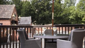 Une table ronde entourée de chaises en osier et surmontée d’un parasol blanc, installée sur une terrasse extérieure avec, en arrière-plan, un bâtiment en briques et des arbres.