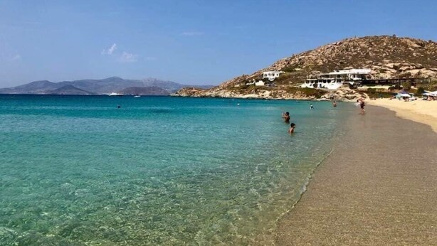 Una spiaggia con acque cristalline turchesi, una costa sabbiosa e una collina rocciosa con edifici sullo sfondo.